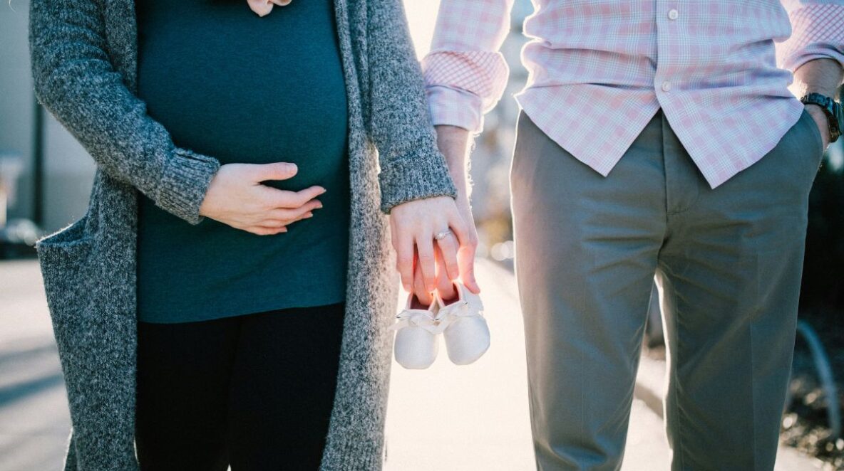 Pregnant woman holding hand with man, both holding baby shoes