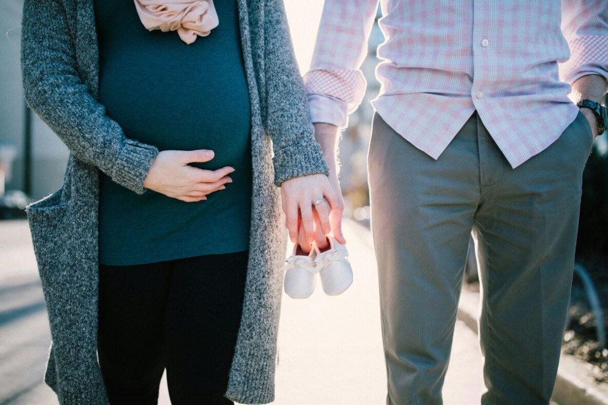Pregnant woman holding hand with man, both holding baby shoes