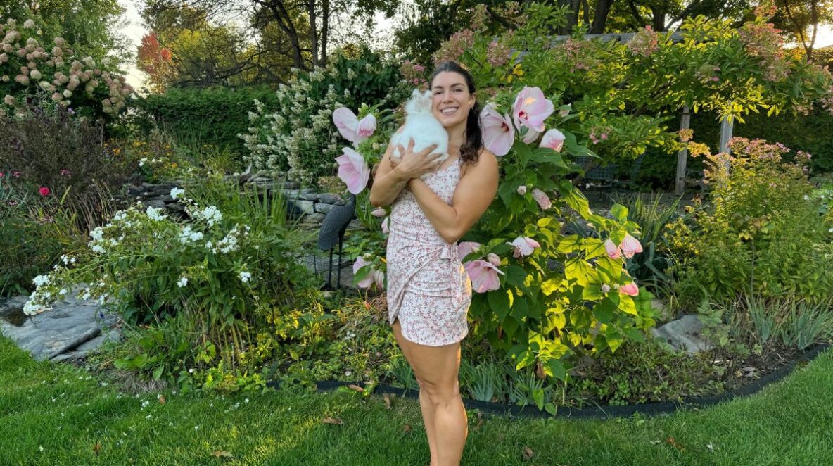 Woman standing and smiling while holding a white bunny rabbit, with a lush green background with flowers.