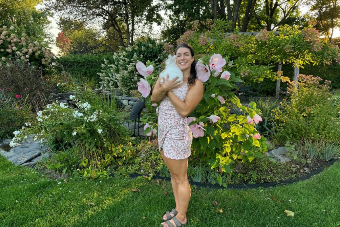 Woman standing and smiling while holding a white bunny rabbit, with a lush green background with flowers.