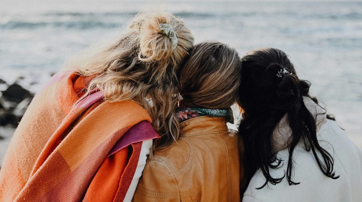 Three women leaning on each other, facing the ocean.