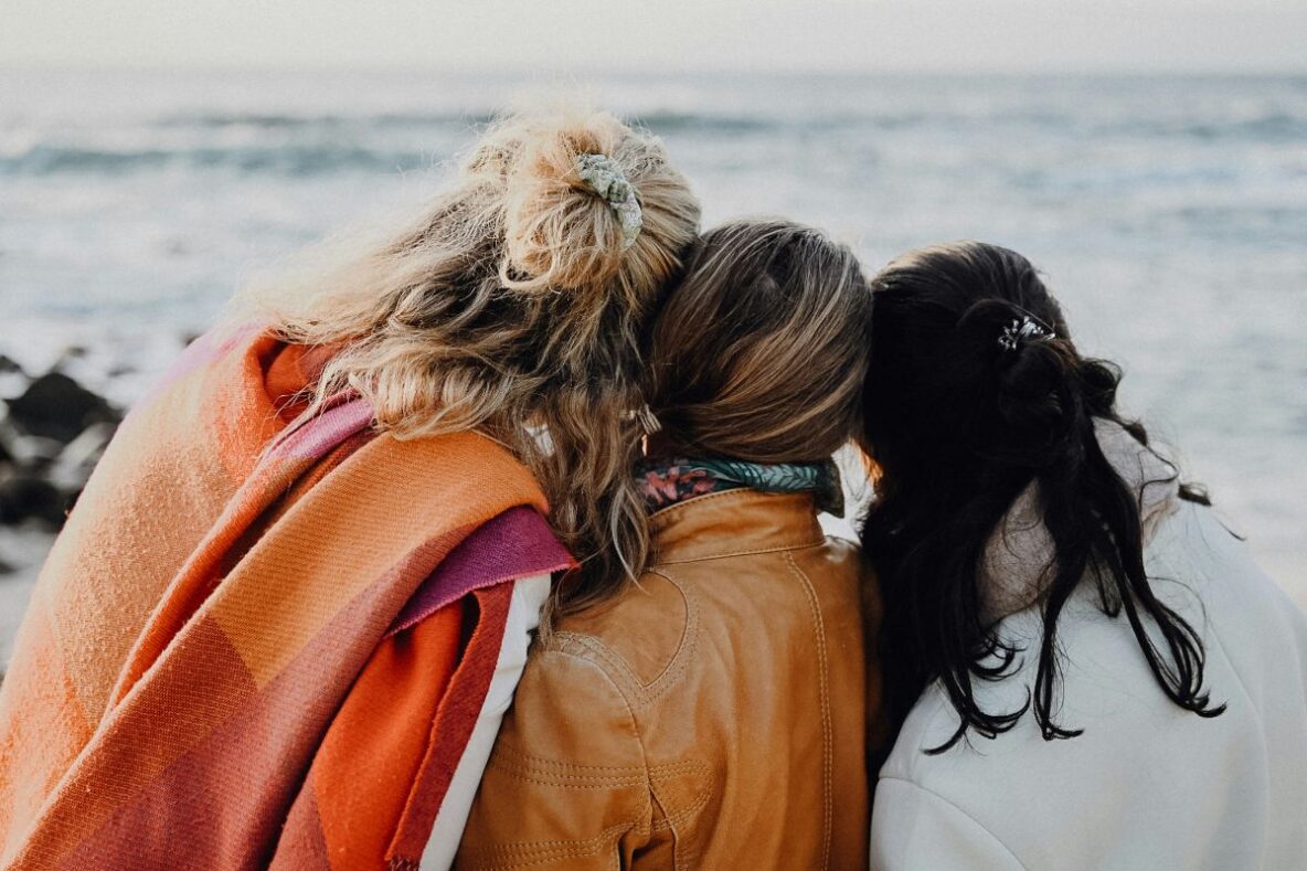 Three women leaning on each other, facing the ocean.